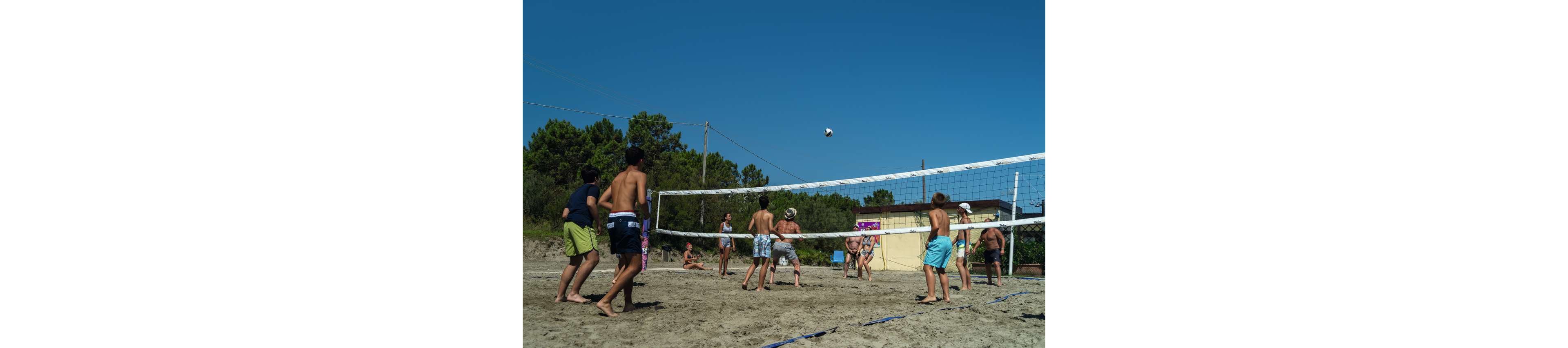A group of young people play volleyball on a sandy outdoor court. The scene takes place on a sunny day with a clear blue sky and trees in the background. Players in swimwear are gathered around the net, watching the ball in midair. Buildings and utility poles are visible behind the court, emphasizing a casual recreational setting.