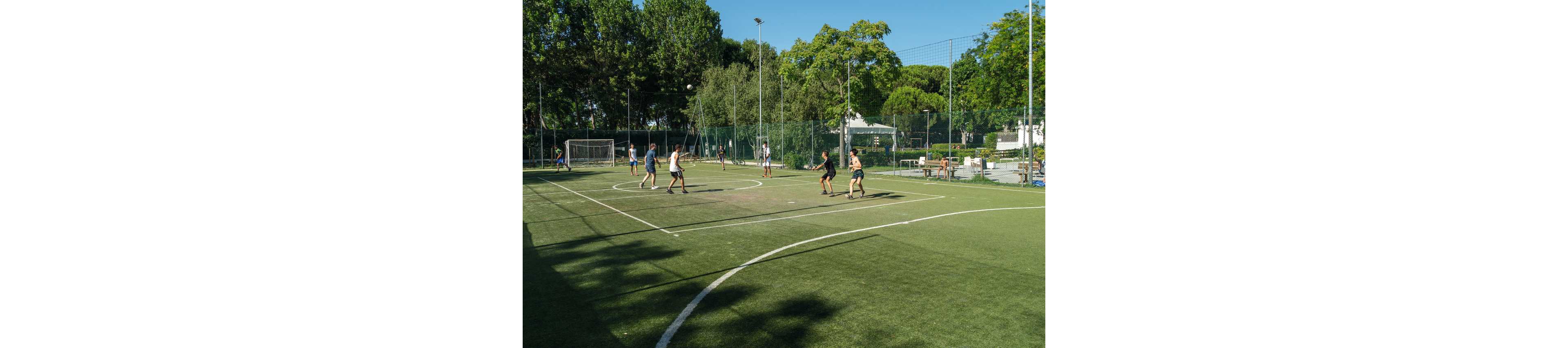 A group of people play a casual soccer game on an outdoor artificial turf field. The scene is set on a sunny day with long shadows and clear blue sky. Tall trees and high chain-link fences surround the pitch, with a goal visible in the background. Spectator benches and sports equipment are placed along the sideline, emphasizing a recreational park environment.