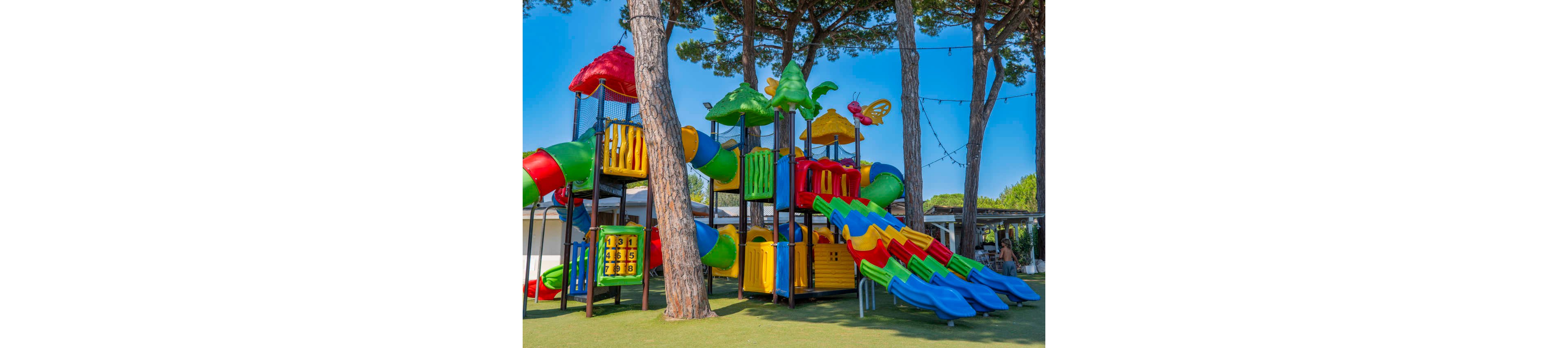 A large, colorful playground structure stands among tall trees in an outdoor park. Multiple bright slides, tunnels, and climbing sections create an inviting play area for children. The equipment features vivid red, yellow, green, and blue elements that contrast with the natural surroundings. In the background, buildings and string lights suggest a recreational or holiday setting.