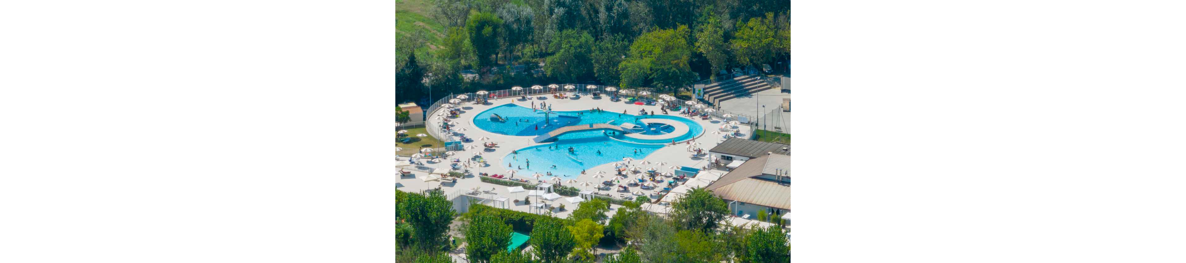 An aerial view shows a large outdoor swimming pool complex surrounded by sun loungers and umbrellas. People are swimming, relaxing on deck chairs, and walking around the pool deck. A curved bridge crosses over the main pool, and smaller circular pools are integrated into the design. The facility is bordered by trees and greenery, giving the scene a resort-like atmosphere on a sunny day.