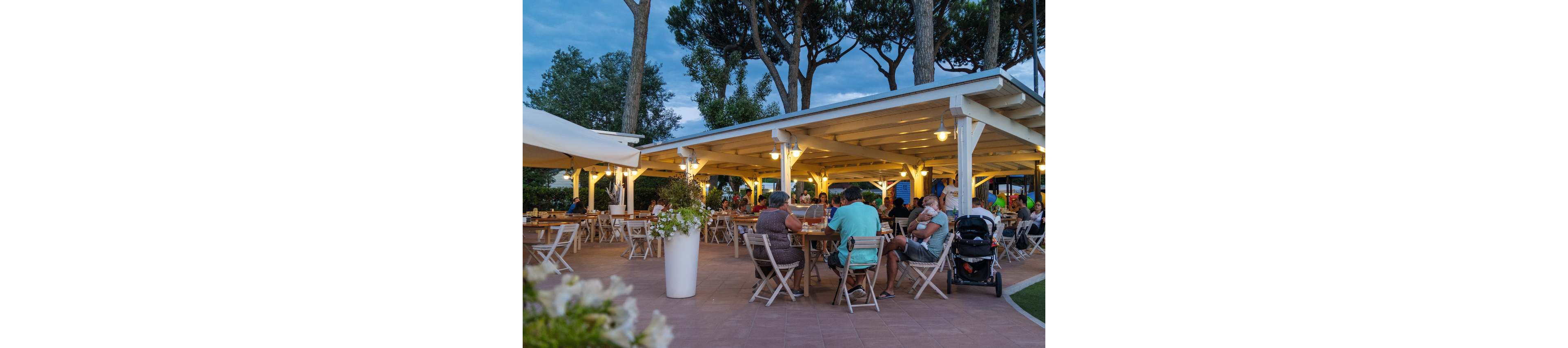 An open-air restaurant terrace is filled with people enjoying an evening meal. Wooden tables and chairs sit beneath a white pergola structure illuminated by warm hanging lights. Families and couples relax, with a stroller visible near one of the tables. Tall trees and a blue evening sky create a calm, vacation-like atmosphere around the dining area.