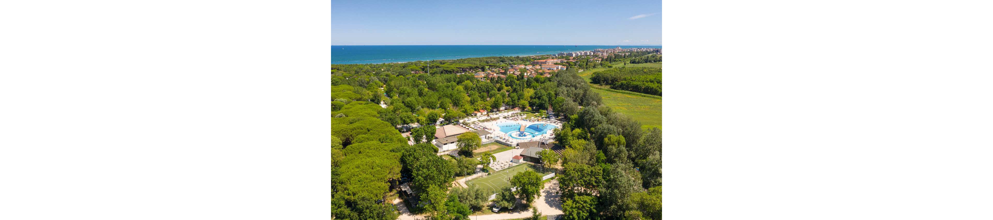 An aerial view shows a large holiday resort nestled within dense green forest near the coastline. At the center of the complex is a bright blue swimming pool surrounded by sun loungers and white buildings. A small sports field is visible in the foreground, while the sea and a distant town stretch across the horizon. The scene suggests a sunny, relaxed vacation atmosphere in a natural setting.
