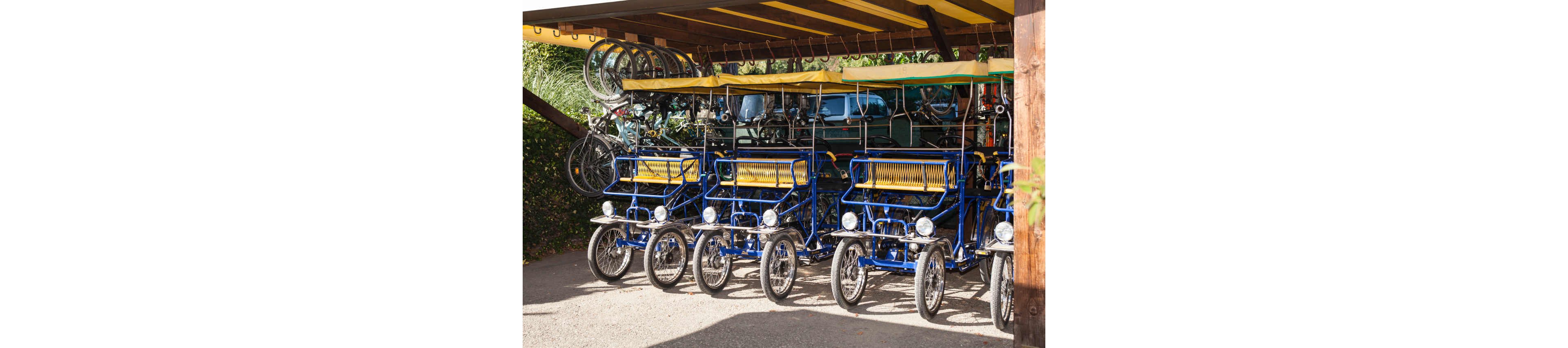Several blue multi-seat pedal carts are parked in a neat row under a wooden shelter. The carts feature yellow seats and canopies, with four wheels and steering wheels visible at the front. Traditional bicycles are stored on racks above the carts, suggesting a rental or leisure facility. Sunlight and surrounding greenery create a bright, inviting outdoor atmosphere.