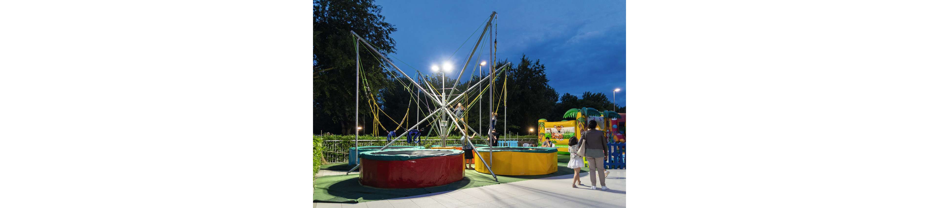 Children play on bungee trampolines in an outdoor amusement area at dusk. The metal structure with elastic cords rises above colorful circular bases in red and yellow. In the background, an inflatable attraction with bright graphics adds to the festive atmosphere. A family walks nearby, illuminated by park lights against a deep blue evening sky.