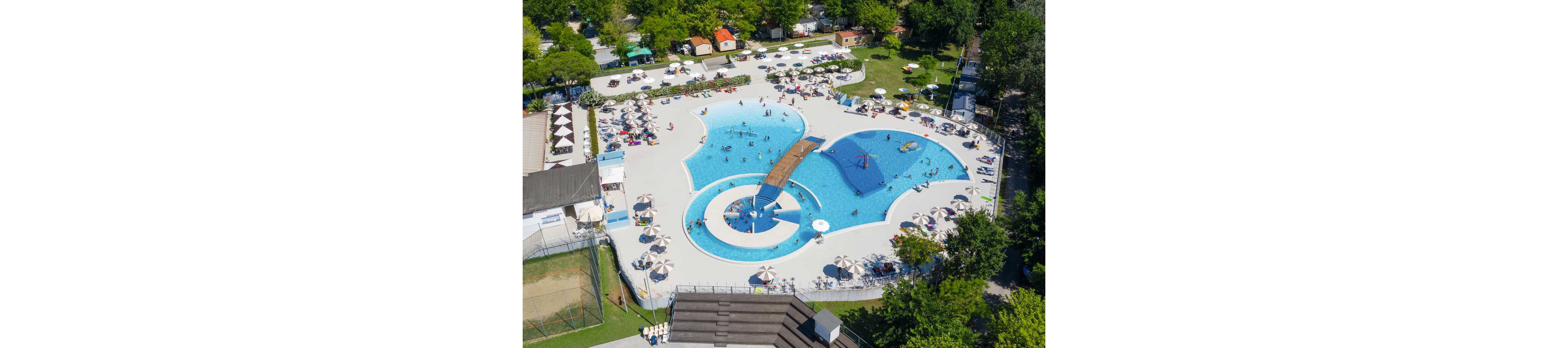 A large outdoor swimming pool complex is seen from above on a sunny day. The pool features curved shapes, shallow areas, and a central bridge with people swimming and wading throughout. Surrounding the water are rows of sun loungers and umbrellas, with trees and small buildings framing the scene. The bright blue water contrasts with the light pool deck, creating a lively vacation atmosphere.