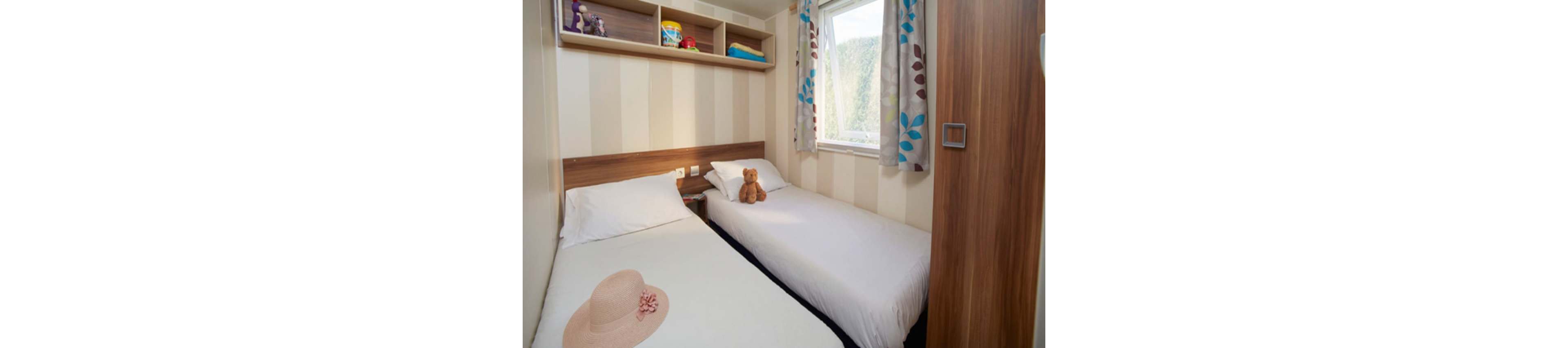 A small, cozy bedroom features two single beds with white linens and simple wooden headboards. A teddy bear sits on one bed while a sun hat rests on the other, adding a personal touch. Built-in shelves above the beds hold a few toys and folded towels. Natural light enters through a window with patterned curtains, brightening the compact, neatly arranged space.