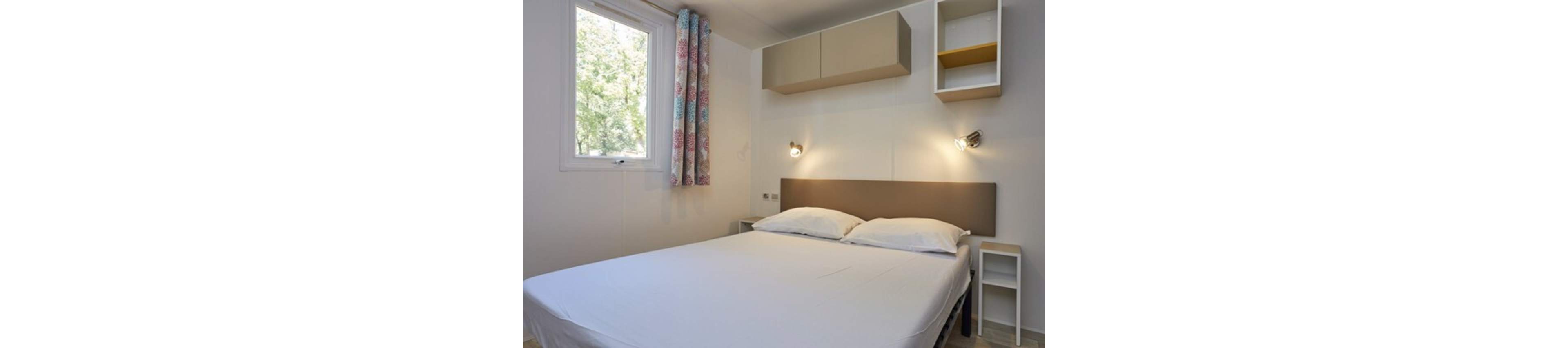 A neatly made double bed with white linens is centered in a small, minimalist bedroom. The room features a padded headboard, two wall-mounted reading lights, and compact bedside tables. Built-in shelves and cabinets provide simple storage above the bed. Natural light enters through a window with patterned curtains, giving the space a clean and airy feel.