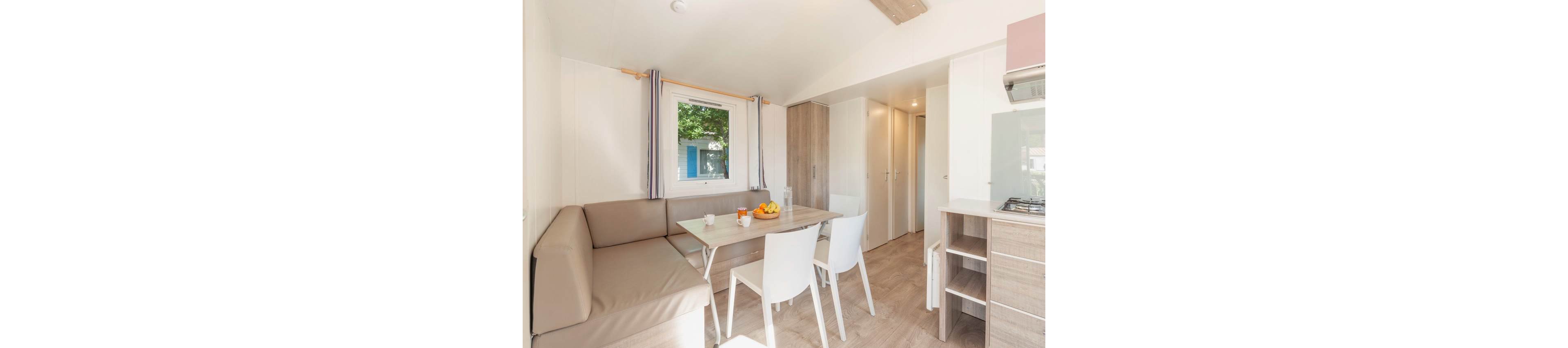 Compact dining and living space inside a modern mobile home. A corner bench and white chairs surround a small table set with cups and a bowl of fruit. Light wood cabinetry, neutral upholstery, and soft natural light create an airy, minimalist feel. A window with striped curtains looks out to greenery, adding a touch of nature to the interior.