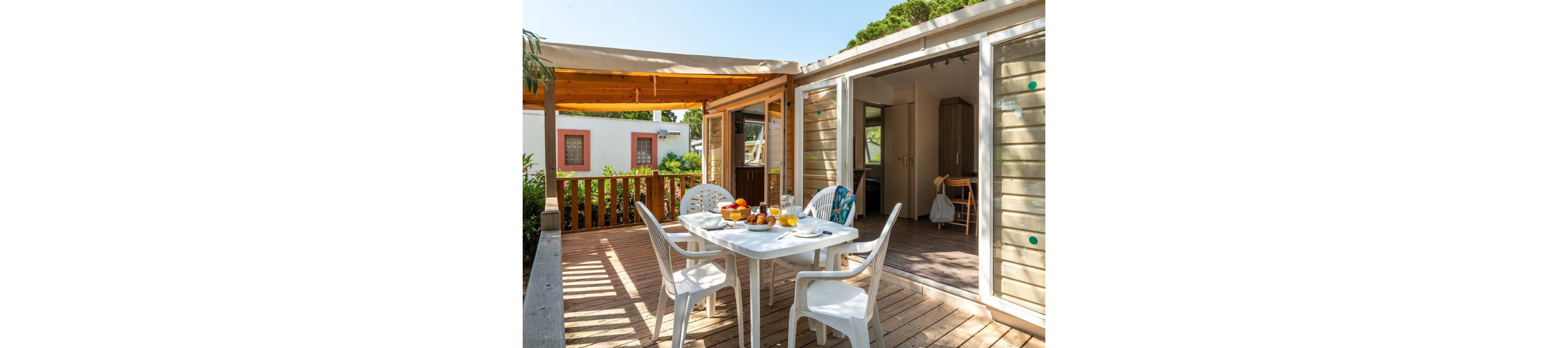 A cozy wooden terrace features a white outdoor dining table set for breakfast with juice, fruit, and pastries. Plastic chairs surround the table under a partial pergola roof, creating a shaded seating area. Sliding glass doors open into a bright cabin interior with simple furnishings. Another small house and lush greenery are visible in the background, suggesting a relaxed holiday setting.