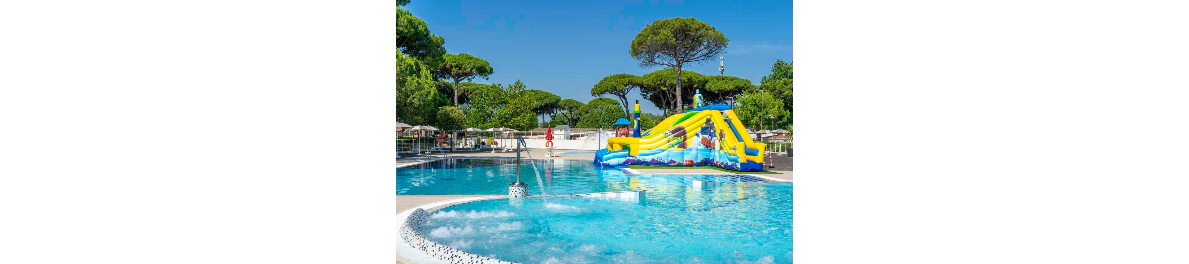 An outdoor swimming pool features a large, colorful inflatable water slide as its main attraction. The pool area is surrounded by tall green trees and sun loungers under umbrellas, suggesting a resort or holiday park setting. Clear blue water, bubbling jets in the foreground, and bright sunshine create a refreshing summer atmosphere. A lifeguard stands near the pool, emphasizing safety in this family-friendly recreational space.