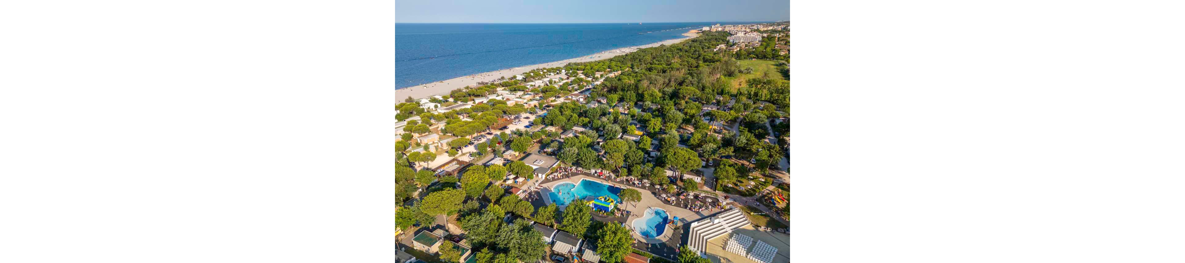 A large seaside holiday resort with pools and campsites stretches along a sandy coastline. The aerial view shows swimming pools, sun loungers, and recreational areas surrounded by dense green trees. Tents, caravans, and small buildings are arranged in neat rows near the beach. The calm blue sea and long shoreline emphasize a relaxed vacation atmosphere.