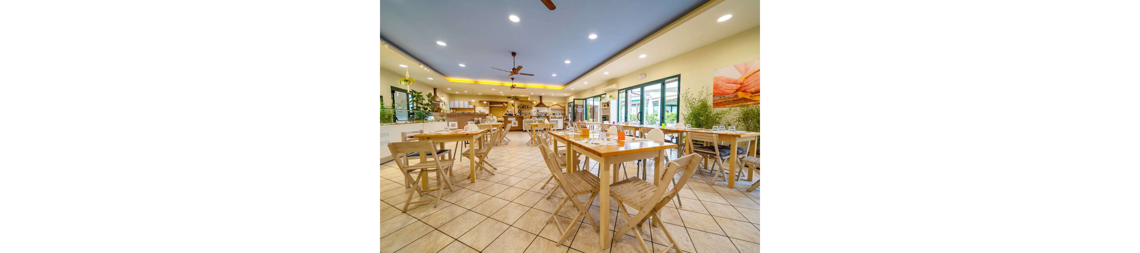 Spacious restaurant interior with light wooden tables and chairs arranged neatly across a tiled floor. Large windows and glass doors line one side, bringing in natural light and views of greenery outside. The ceiling features recessed lighting and ceiling fans, creating a comfortable and airy atmosphere. Table settings with glasses and condiments suggest the space is ready to welcome guests for a meal.