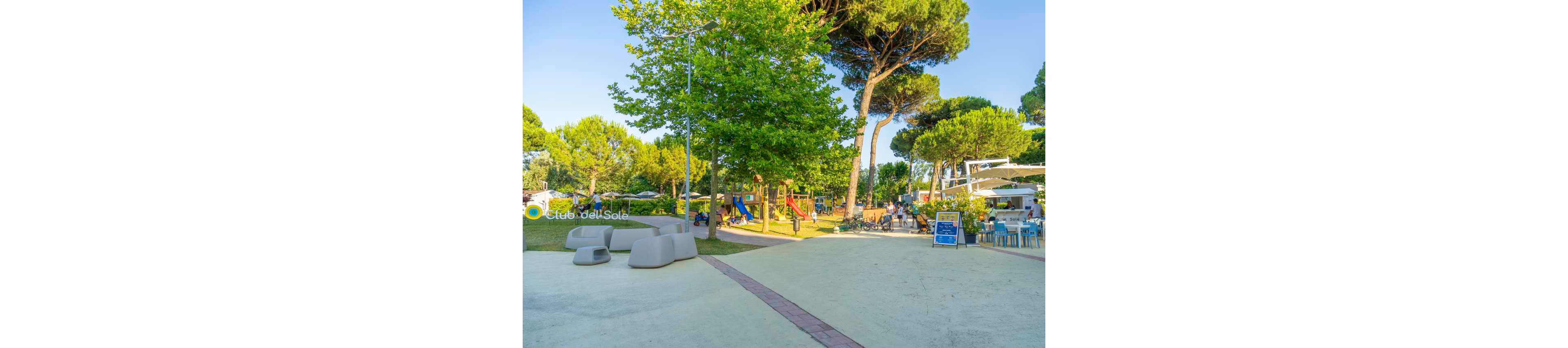 An outdoor leisure area features a modern playground surrounded by tall green trees on a sunny day. Families and children are visible near play structures and seating areas, creating a lively vacation atmosphere. To the right, an open-air café or bar with tables and chairs hosts relaxed visitors. Bright colors, clear blue sky, and soft paved surfaces contribute to a cheerful, summery mood. The text "Club del Sole" appears on the left side of the image.