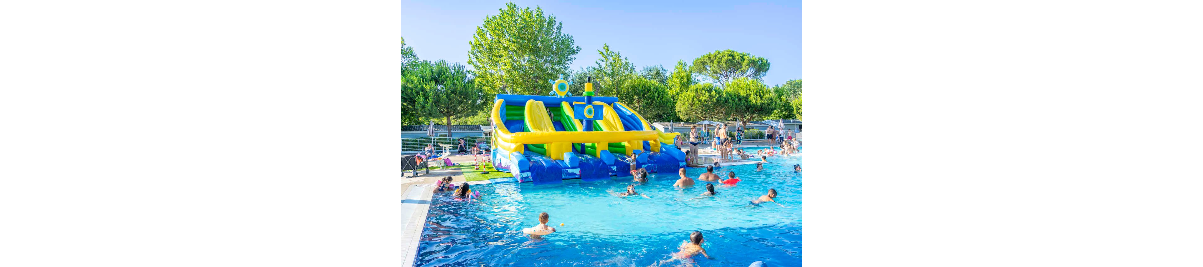 A large outdoor swimming pool is filled with people enjoying a sunny day. At the far end of the pool, a colorful inflatable water slide structure with multiple lanes stands out. Children and adults play in the water while others wait near the slide and relax around the pool deck. Lush green trees and clear blue skies create a bright, cheerful summer atmosphere.