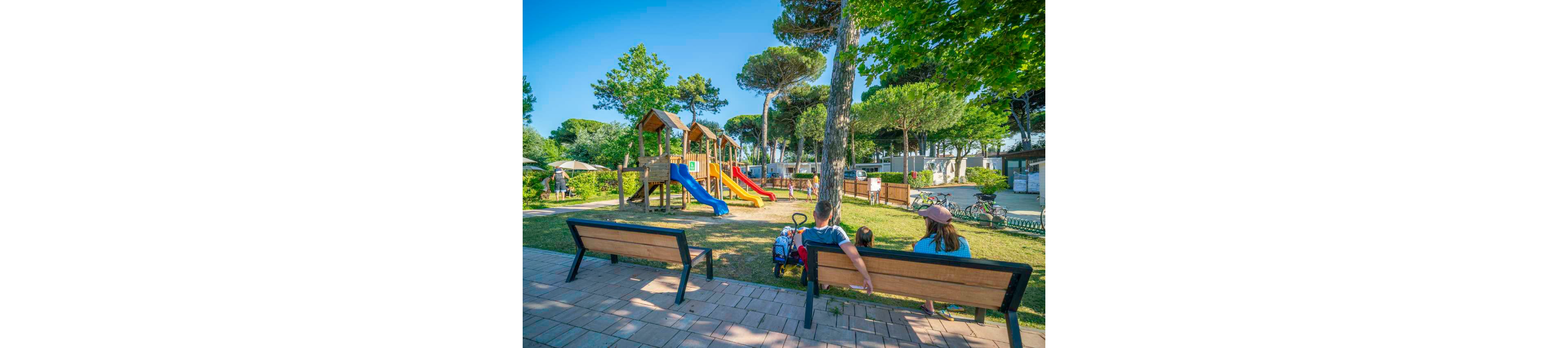 A family sits on a bench watching children play at a colorful outdoor playground. The playground features wooden climbing structures with bright red, yellow, and blue slides. Tall green trees provide shade and create a pleasant park atmosphere. Other people walk and relax in the background, suggesting a friendly community or holiday campsite setting.