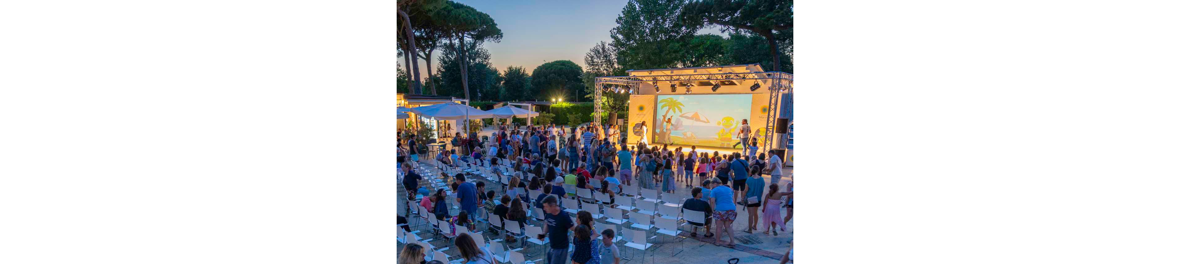 An outdoor stage hosts a colorful family show at sunset, featuring a large screen with a beach and cartoon characters. A crowd of adults and children gathers in front of the stage while others sit or stand among rows of white chairs. Food stalls and lit canopies are visible to the side, adding to the festive atmosphere. Tall trees frame the open-air venue under a clear evening sky.