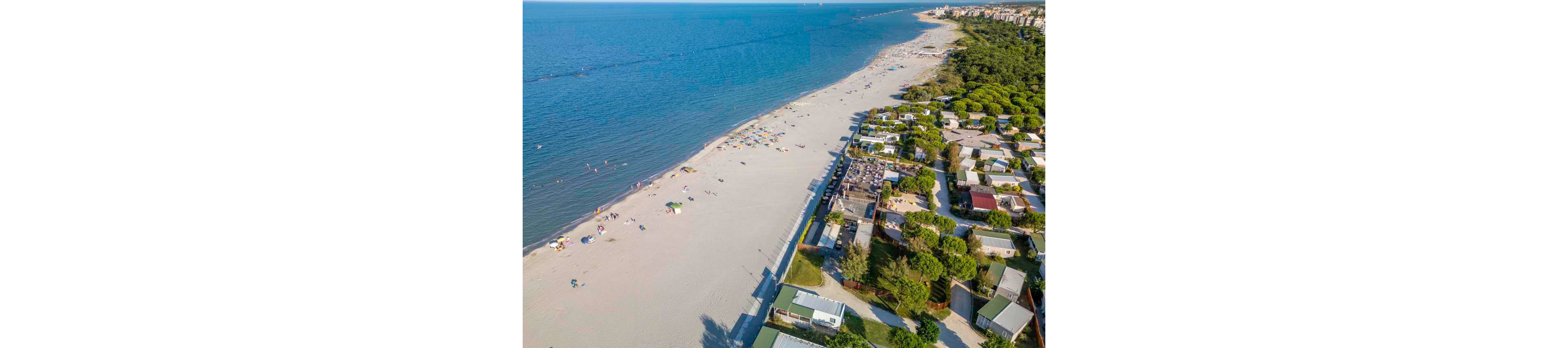 A wide sandy beach stretches along a calm blue sea, viewed from above. On one side of the image, a coastal campground with cabins and trees lines the shore. Sunbathers and swimmers are scattered across the sand and in the shallow water, with colorful umbrellas providing shade. The scene suggests a relaxed summer holiday atmosphere in a popular seaside destination.