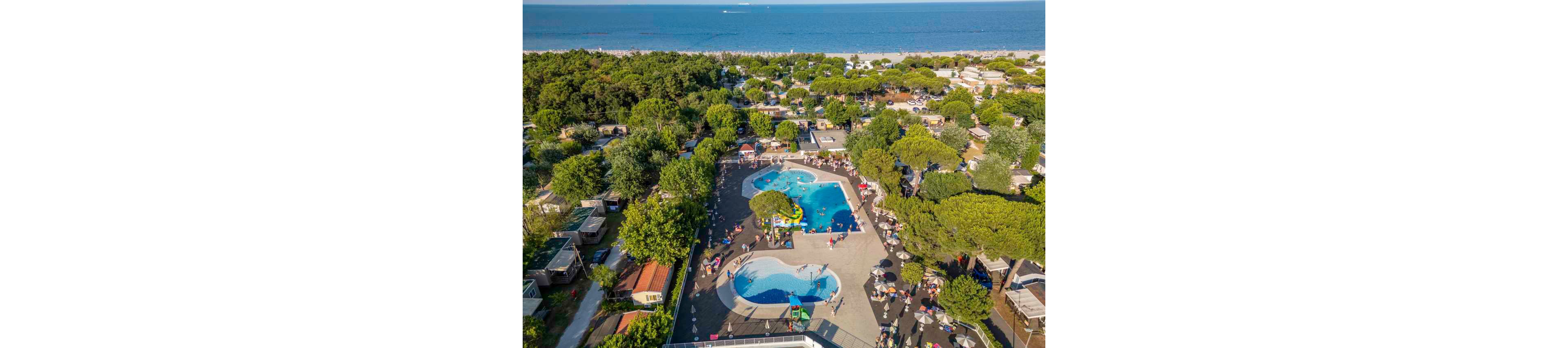 A large outdoor swimming pool complex is captured from above within a lush seaside campsite. Multiple pools with blue water are surrounded by sun loungers, umbrellas, and people enjoying the warm weather. Trees and mobile homes fill the campground, creating a green contrast against the paved pool area. In the distance, a sandy beach and calm sea stretch across the horizon, emphasizing the holiday atmosphere.