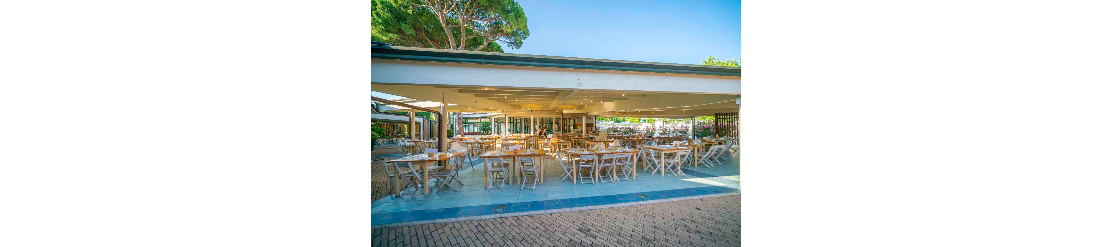 An open-air restaurant features neatly arranged wooden tables and chairs under a wide, covered terrace. The space is bright and inviting, with natural light streaming in from the open sides. Green trees and flowering bushes surround the dining area, suggesting a coastal or resort setting. The restaurant appears ready for guests, with table settings prepared but no diners yet seated.