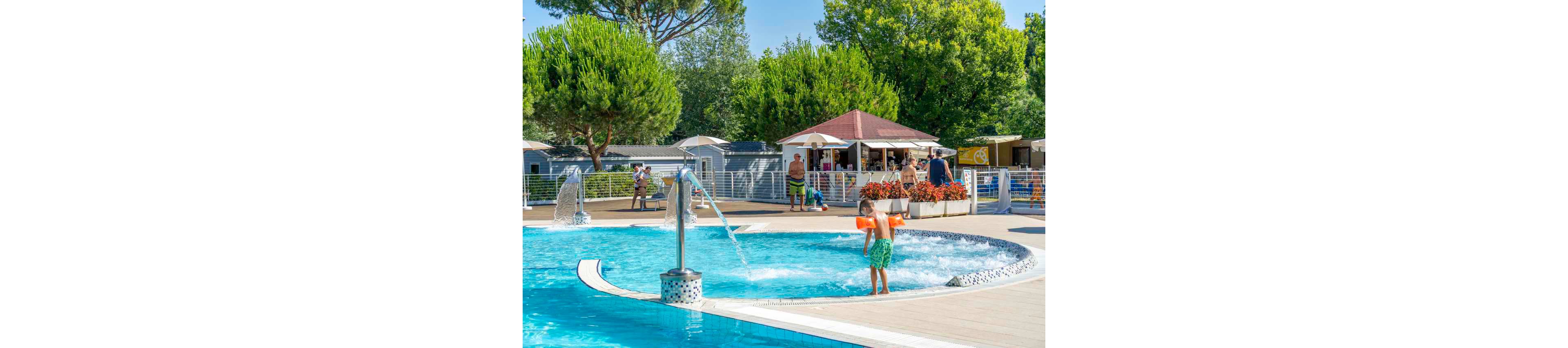 A child wearing orange floaties plays in a shallow outdoor swimming pool with water features. Families and children relax around the pool deck under umbrellas near a small poolside bar or kiosk. Tall green trees and bright flowers surround the area, creating a lush, summery atmosphere. The clear blue water and sunny sky suggest a warm vacation setting at a resort or campground.