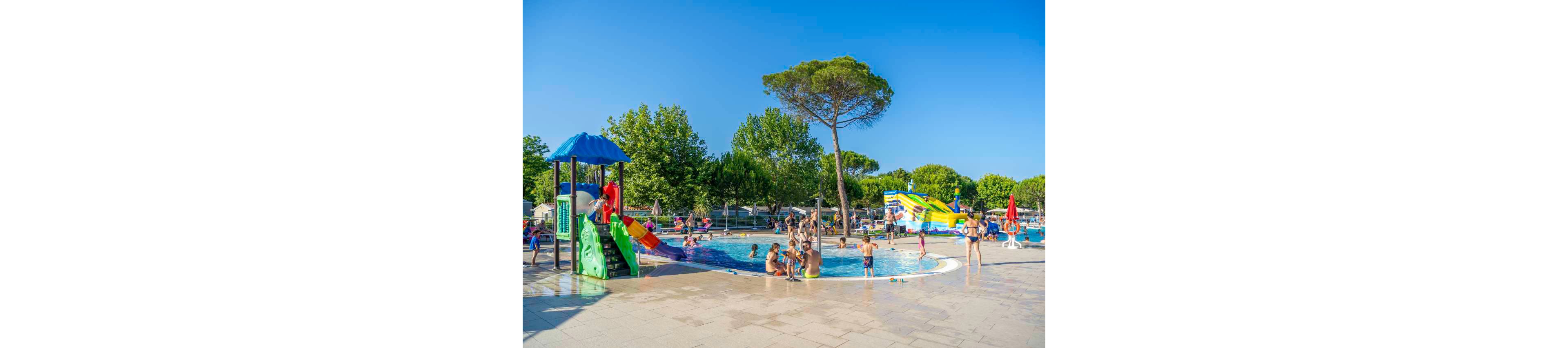 Children and adults play together in a shallow outdoor swimming pool at a family waterpark. Colorful slides and water features surround the pool, creating a fun and lively atmosphere. Tall trees and green foliage frame the scene under a clear blue sky. People relax, splash, and socialize, suggesting a warm summer holiday setting.
