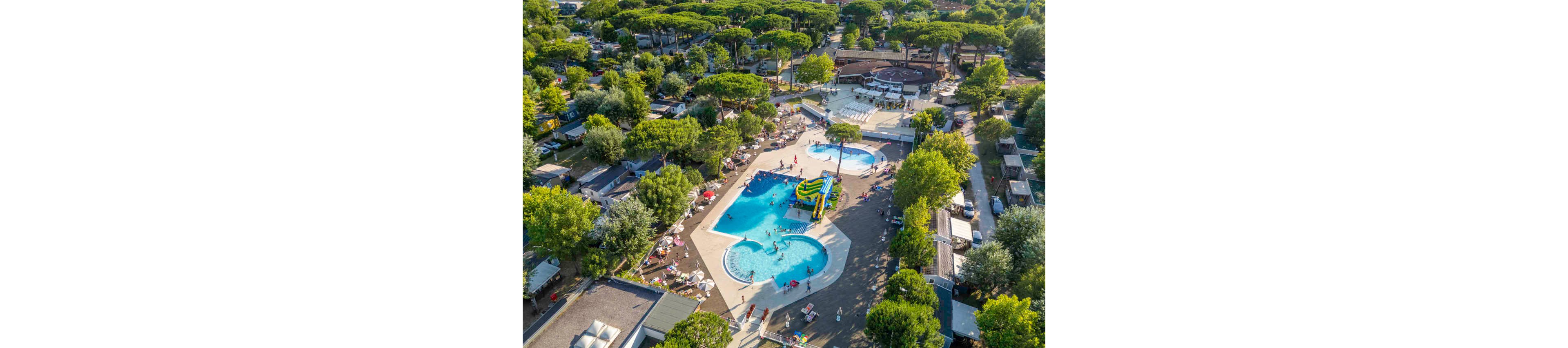 A large outdoor pool complex is seen from above in the middle of a green, tree-filled resort. Several swimming pools of different shapes are surrounded by sun loungers, umbrellas, and small buildings. People of various ages enjoy the water, slides, and poolside areas, creating a lively holiday atmosphere. The surrounding pine trees and cabins suggest a campground or vacation village setting.