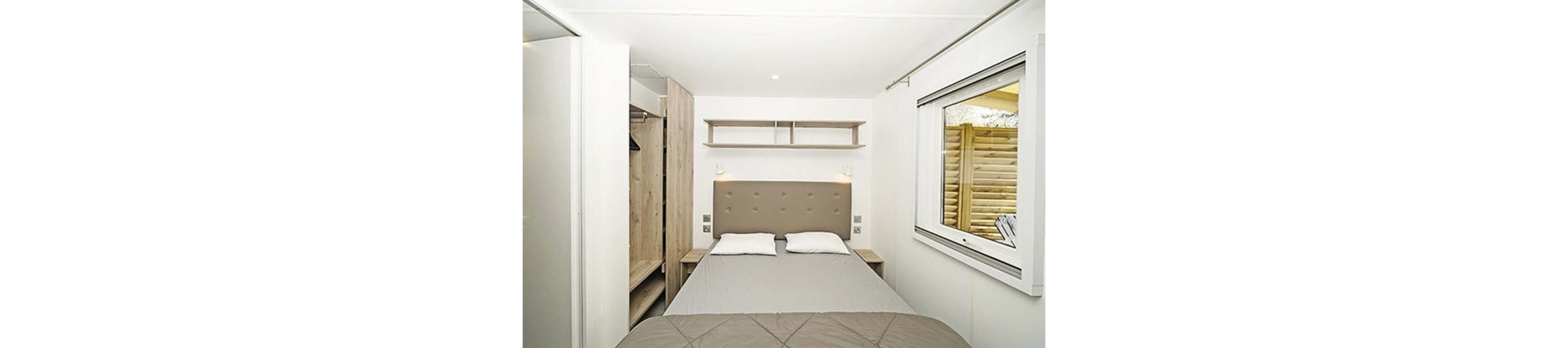 A compact bedroom features a neatly made double bed with a padded beige headboard and two white pillows. Built-in wooden wardrobes and open shelving provide storage on the left side of the room. A large window on the right lets in natural light and offers a view of a wooden privacy screen outside. The decor is minimalist with neutral tones, creating a clean and modern atmosphere.