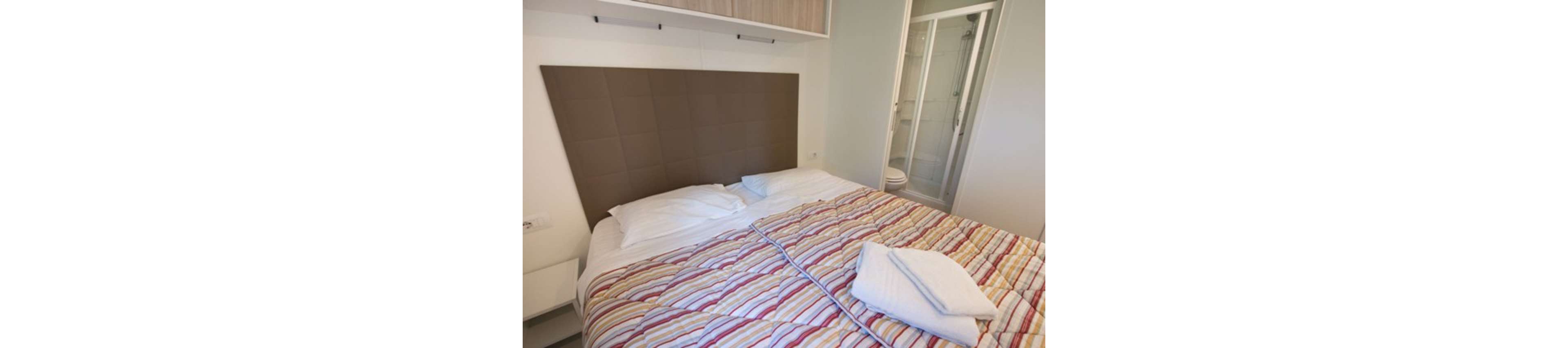 A neatly made double bed with colorful striped bedding dominates a compact modern bedroom. Soft pillows and folded white towels rest on top of the bed, suggesting a prepared guest space. The room features a large padded headboard, built-in storage above the bed, and neutral-colored walls. A glass shower enclosure is visible through an open door, indicating an en-suite bathroom in this contemporary accommodation.