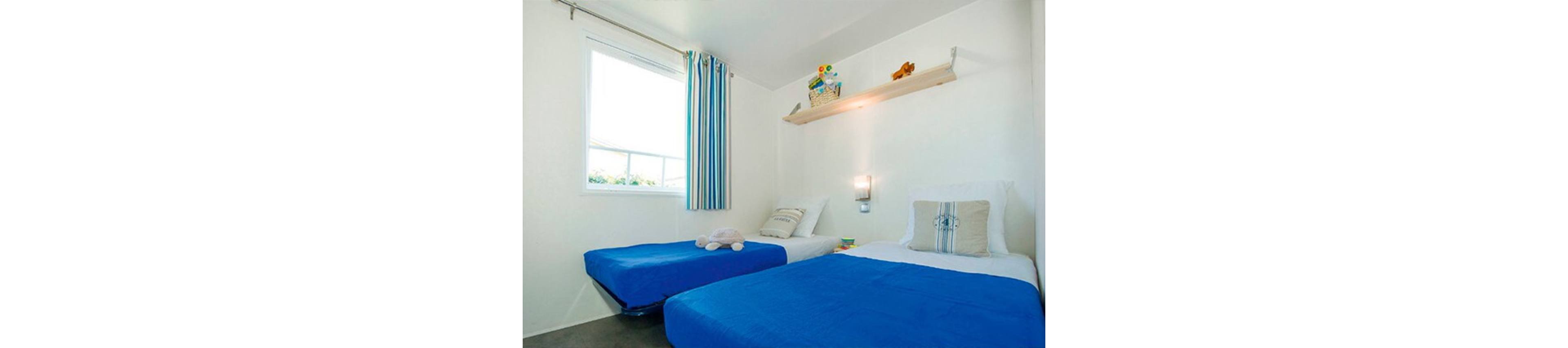 A small, bright bedroom features two twin beds with vivid blue bedspreads and white sheets. One bed has a plush toy placed near the pillow, adding a playful touch. A wall-mounted shelf above the beds holds a few colorful decorative items and a basket. Natural light enters through a window with striped blue and white curtains, while a wall sconce provides additional lighting on the white walls.