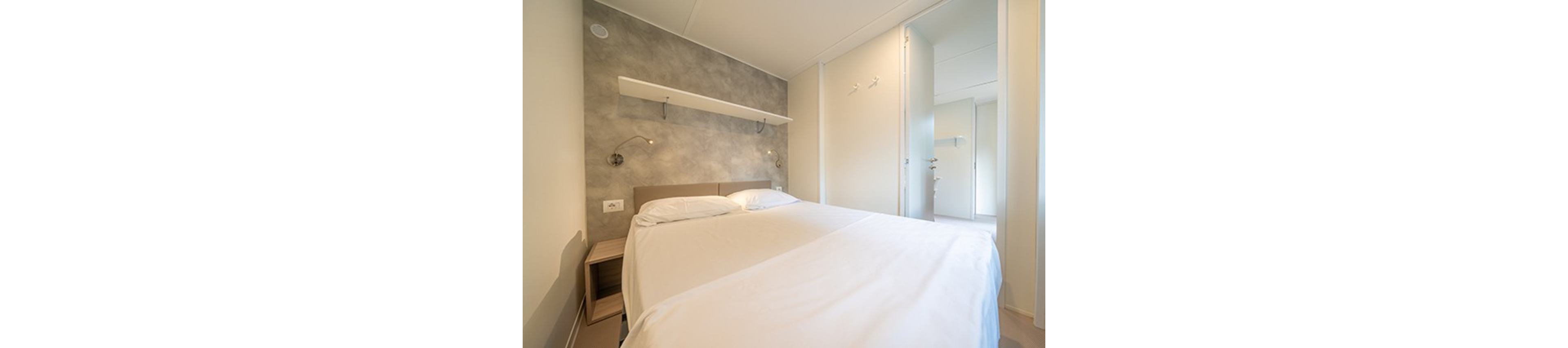 A clean, modern bedroom features a neatly made double bed with white linens. The room has light-colored walls, a textured accent wall, and built-in reading lights above the headboard. A simple floating shelf and small bedside table add to the minimalist decor. An open doorway reveals an adjacent bright hallway or room, enhancing the spacious feel.