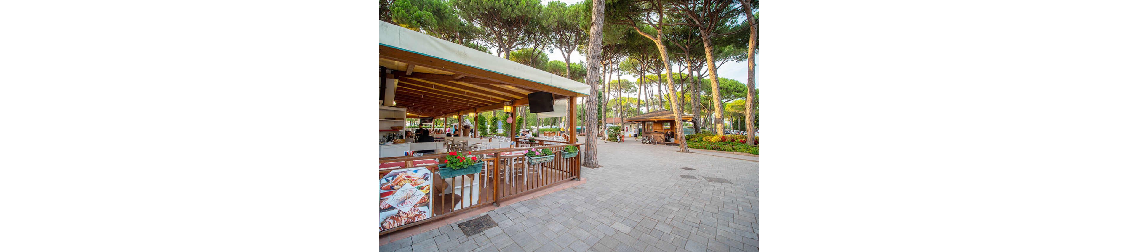An open-air café with wooden beams and white seating sits on the edge of a paved plaza surrounded by tall pine trees. Flower boxes with red blooms decorate the railing, and a food poster hangs near the entrance. A few people are visible inside the café, while another small kiosk stands further down the walkway. The scene is bright and inviting, suggesting a relaxed outdoor dining atmosphere in a park-like setting.