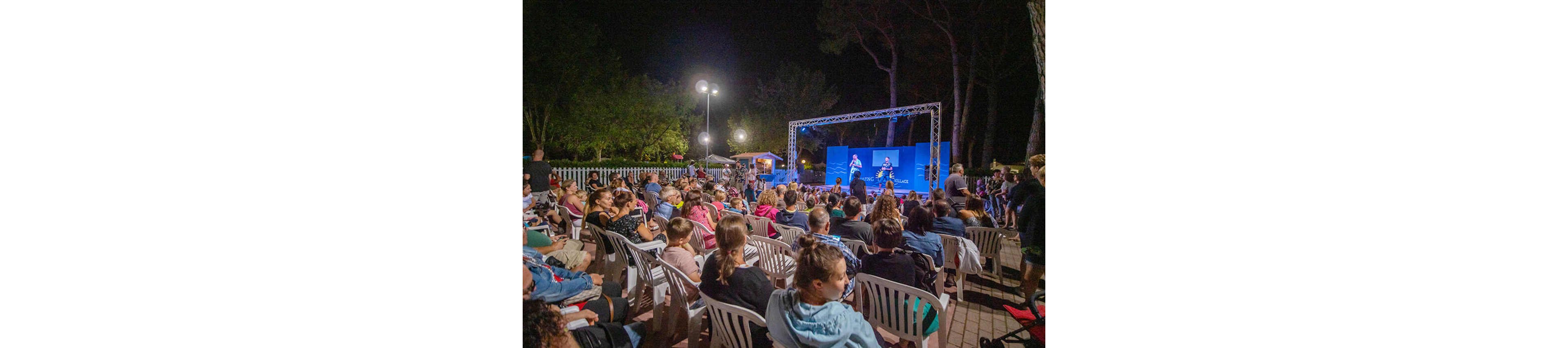A large audience sits on plastic chairs watching a live performance on an outdoor stage at night. Two performers stand under bright stage lights against a blue backdrop, framed by a metal truss structure. The crowd includes adults and children, all facing the stage in a relaxed, informal setting. Trees and small buildings are visible in the background, adding to the open-air atmosphere.