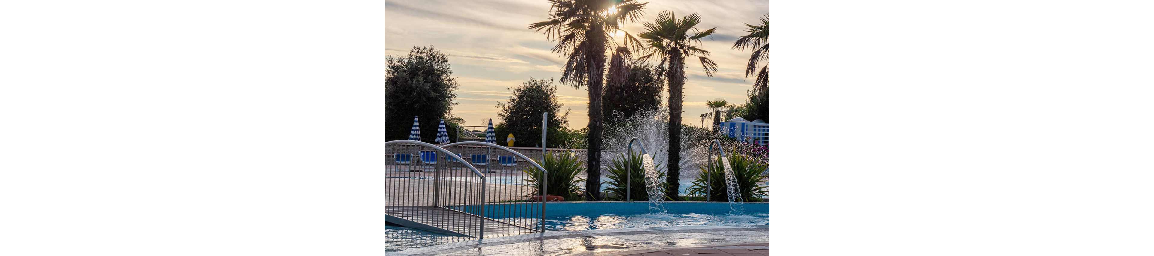 An outdoor resort swimming pool features arched water fountains spraying into the blue water. Palm trees and lush greenery surround the pool, creating a tropical atmosphere. A metal bridge crosses part of the pool, and striped umbrellas and lounge chairs are visible in the background. The sun is low in the sky, casting warm light and long shadows across the scene.