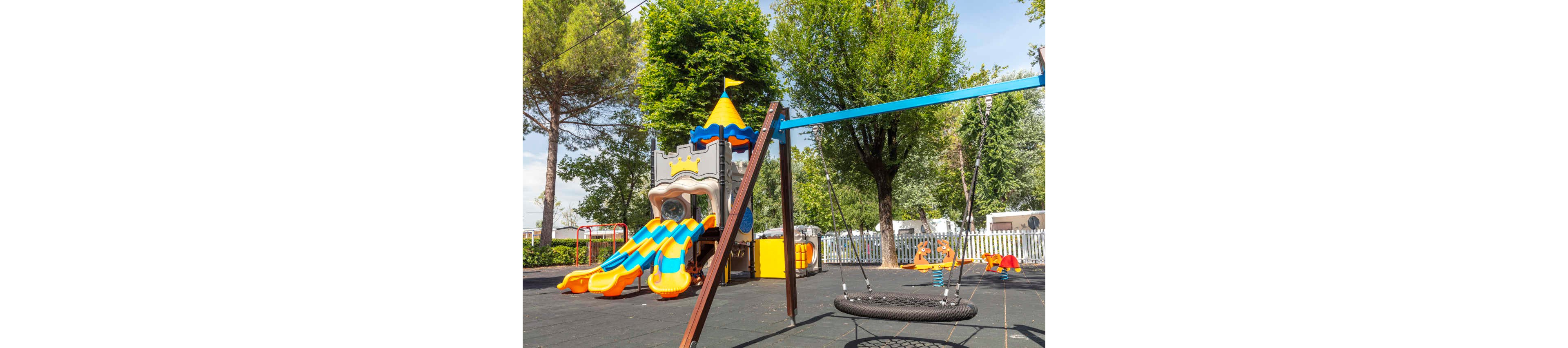 A bright playground features a castle-themed play structure with multiple orange and blue slides. In the foreground, a circular net swing hangs from a sturdy metal frame. The area is surfaced with safety tiles and surrounded by trees and greenery. A white fence and additional play equipment are visible in the background, suggesting a family-friendly park setting.