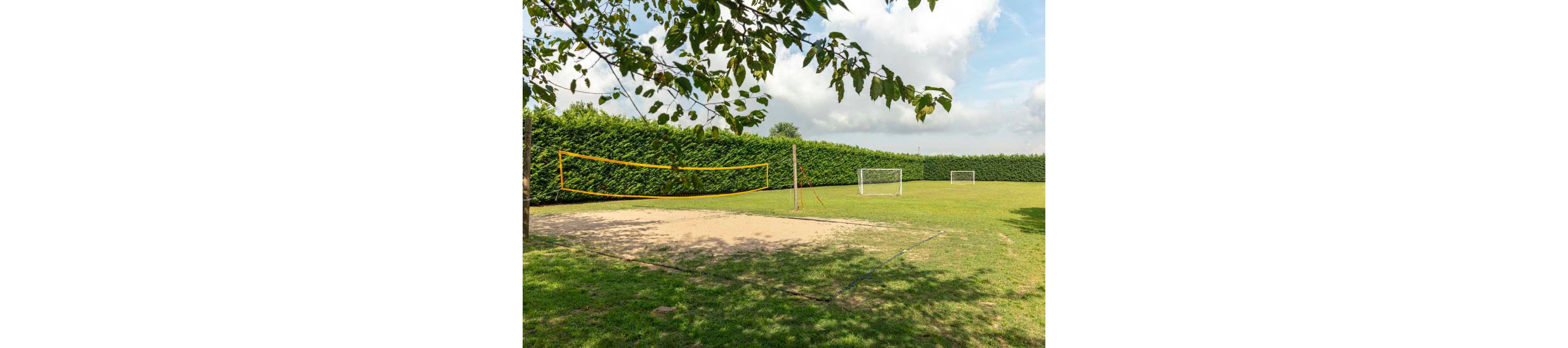 An outdoor sports area features a sand volleyball court with a bright yellow net. The court is set within a large grassy field bordered by tall green hedges. Two small soccer goals stand on the grass behind the volleyball court. A tree branch frames the scene in the foreground under a partly cloudy sky. The setting feels peaceful and recreational, ideal for casual games and outdoor activities.