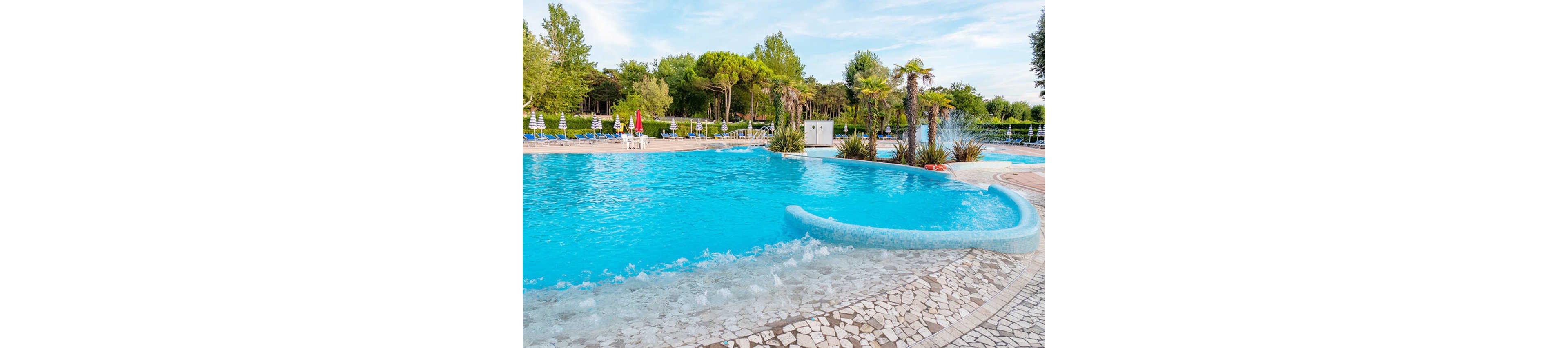 A large, bright blue swimming pool is surrounded by lush greenery and palm trees. The pool features a curved edge with gentle waves and a shallow entry area. In the background, rows of sun loungers and umbrellas line the deck, suggesting a relaxing resort setting. The scene is calm and inviting under a clear sky, ideal for leisure and vacation time.