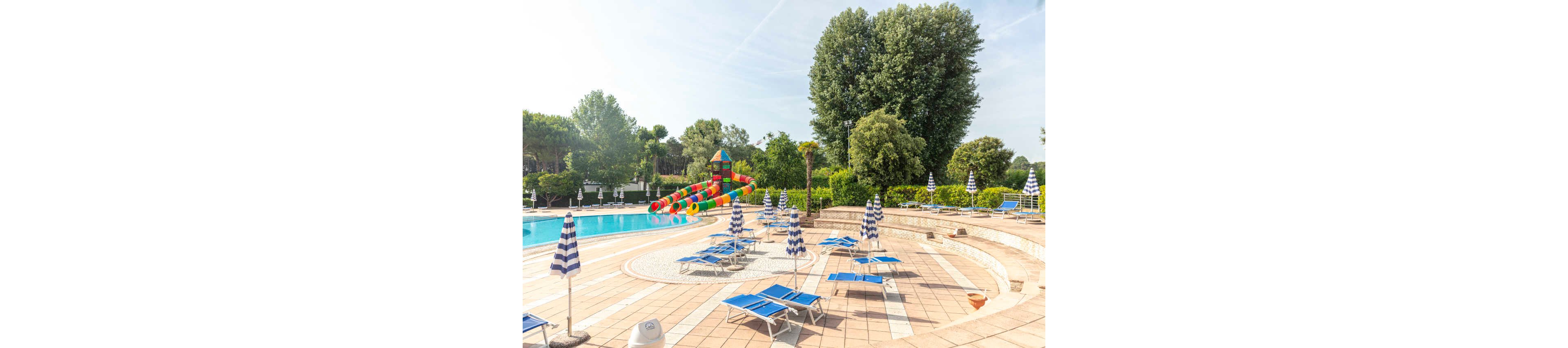 An outdoor resort swimming pool is surrounded by blue lounge chairs and striped umbrellas on a tiled deck. In the background, a colorful inflatable water slide structure rises near the pool edge. Tall green trees and hedges frame the scene under a bright, clear sky. The area appears calm and empty, suggesting an early morning or off-peak time at the pool.