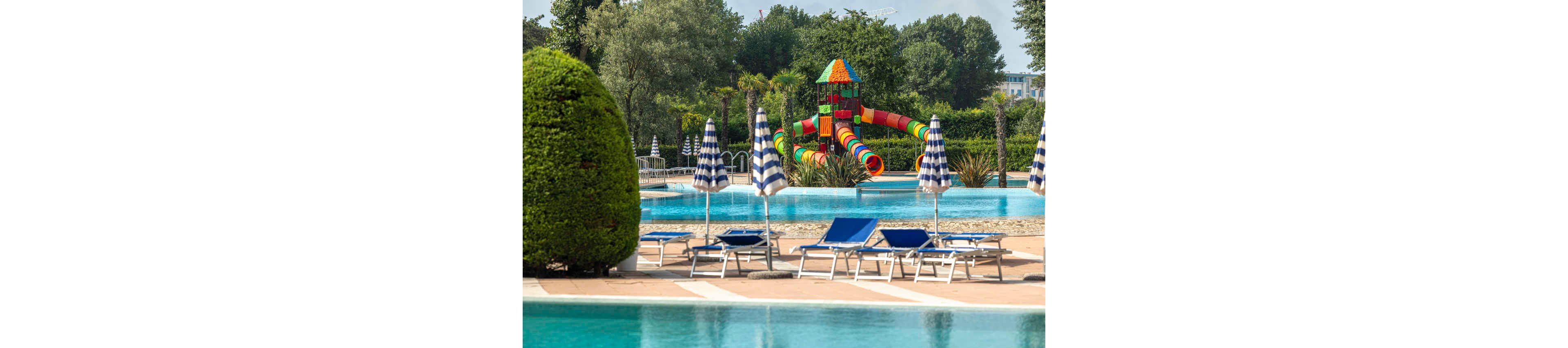 An outdoor swimming pool complex features multiple clear blue pools surrounded by sun loungers and striped umbrellas. In the background, a large colorful playground-style water slide structure stands out with bright red, yellow, green, and blue tubes. Lush greenery and manicured bushes frame the scene, creating a resort-like atmosphere. The area appears calm and empty, suggesting an early or off-peak time at the pool.
