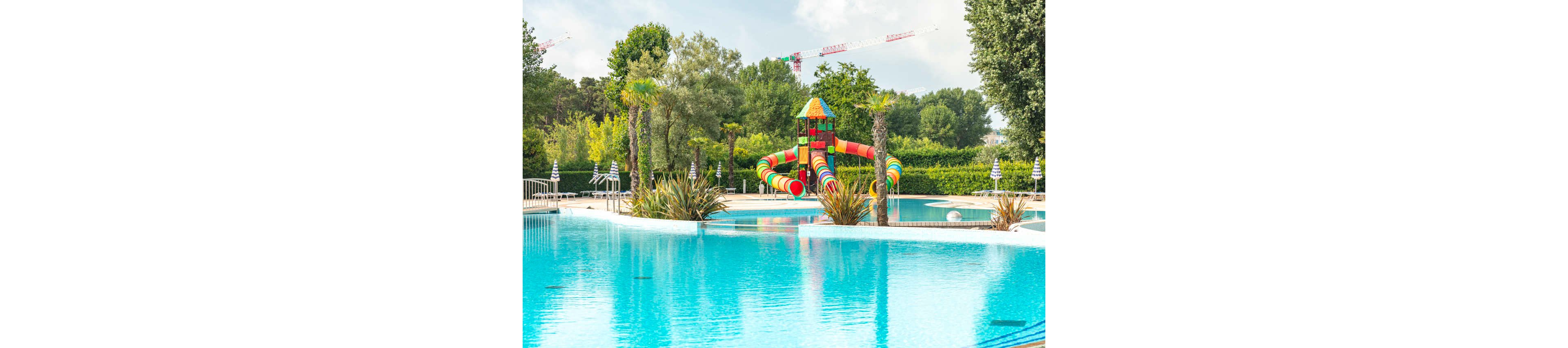 A bright outdoor swimming pool features a colorful multi-tube waterslide structure in the background. The pool is surrounded by lush green trees, hedges, and a few palm-like plants near the water’s edge. Striped umbrellas and metal railings line the perimeter, suggesting a family-friendly resort or water park setting. The calm blue water reflects the vibrant slides and clear sky, creating a cheerful summer atmosphere.