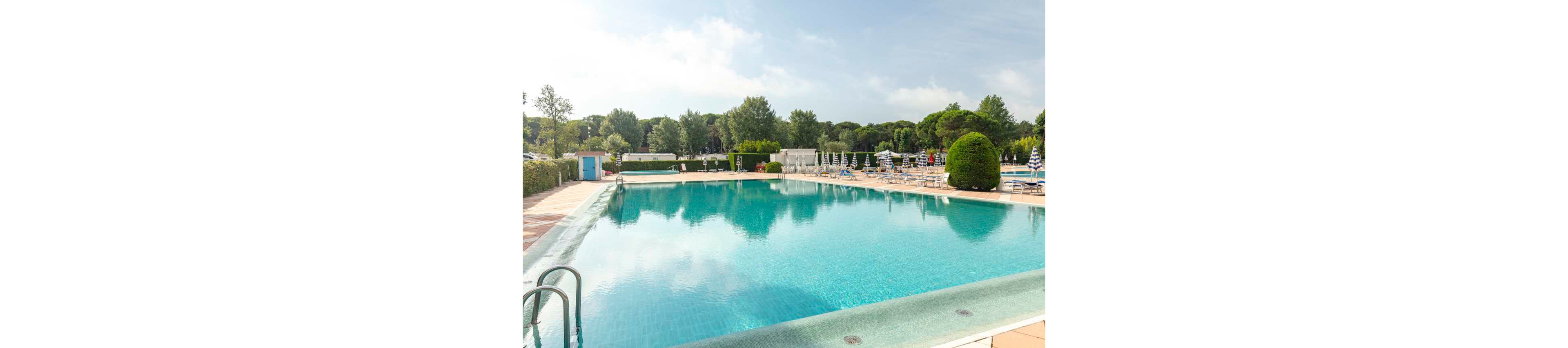 A large outdoor swimming pool glistens under a bright sky in a resort or recreational area. Sun loungers and striped umbrellas line the poolside, suggesting a relaxing leisure setting. Trees and neatly trimmed hedges surround the pool, creating a green, tranquil backdrop. The overall scene feels calm, inviting, and ideal for summer relaxation.