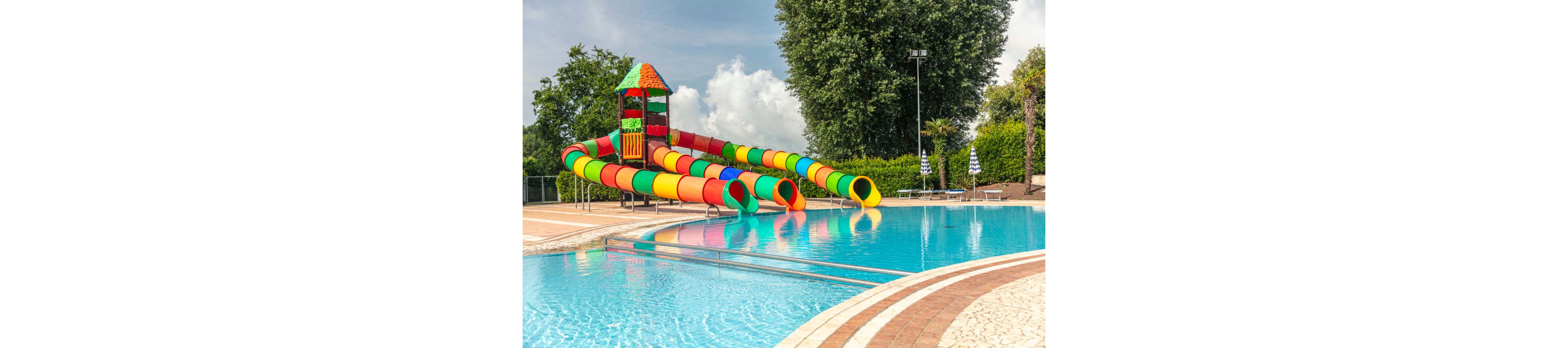 A brightly colored set of tubular water slides leads into a clear blue swimming pool. The slides are red, yellow, green, and orange, attached to a small tower structure. The scene is outdoors on a sunny day with lush green trees and hedges in the background. Poolside paving and metal railings frame the water, suggesting a family-friendly recreational area.