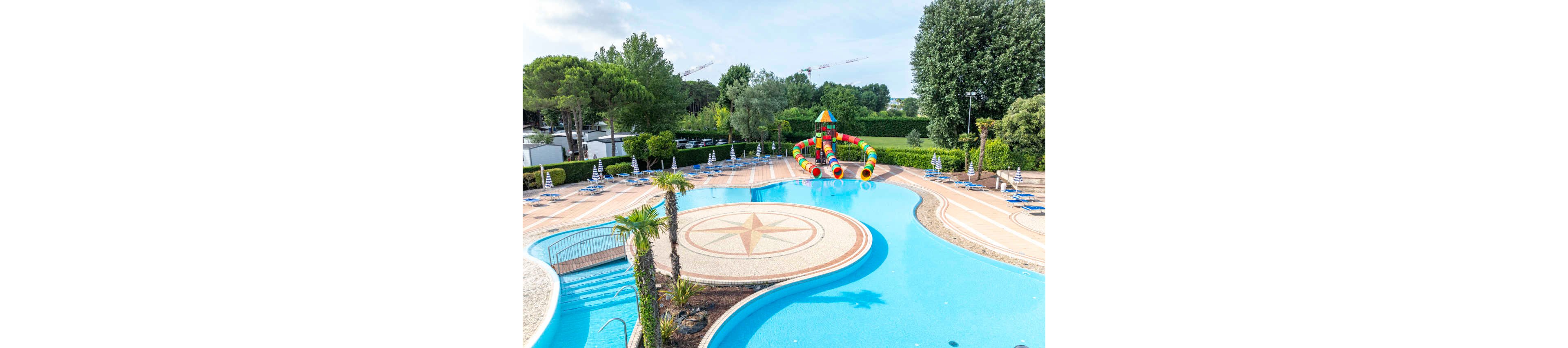A large outdoor swimming pool features a central circular area with a compass-style star design in the tile. Surrounding the pool are numerous lounge chairs and umbrellas neatly arranged on the deck. At one end of the pool, a bright multicolored inflatable water slide structure stands out, designed for children’s play. Lush green trees and hedges enclose the area, creating a private resort-like atmosphere under a clear sky.