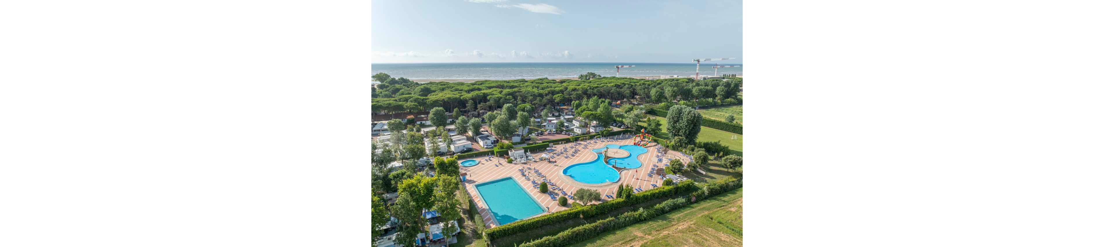 An aerial view shows a large campsite with multiple swimming pools near a coastline. The central area features a freeform pool complex with sun loungers neatly arranged around it. A rectangular lap pool sits beside the main pool, and a small water play area with slides is visible. The campsite is surrounded by trees and greenery, with the sea and sandy beach stretching out in the background.