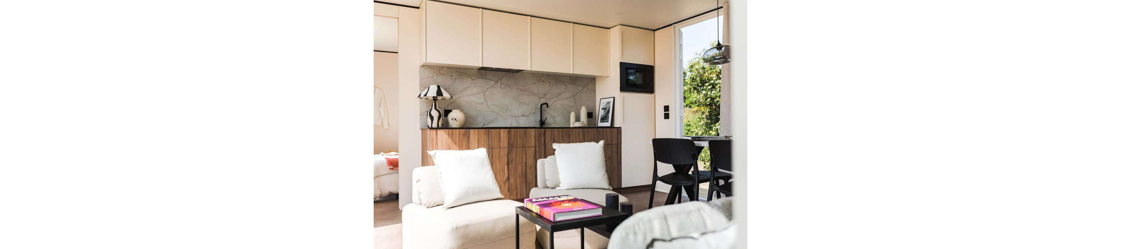 A contemporary open-plan living space features a compact kitchen with light upper cabinets and wood-grain lower cabinetry. A marble-look backsplash and minimalist black faucet add a refined touch to the small kitchen area. In the foreground, a cozy seating area with white cushions faces a low black coffee table holding colorful books. Large windows and an open doorway bring in natural light and views of greenery outside, creating an airy, relaxed atmosphere.