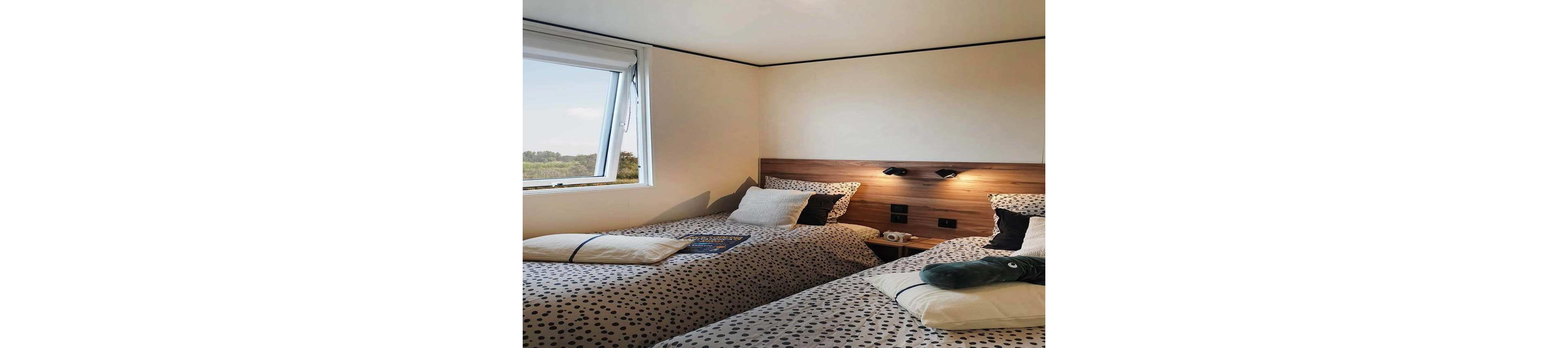 A compact bedroom features two twin beds with matching polka dot bedding and accent pillows. A large window on the left lets in natural light and offers a view of trees and open landscape. The wooden headboard wall includes built-in reading lights and power outlets above a small bedside table. A plush toy and a magazine on the beds add a relaxed, homely touch to the modern interior.