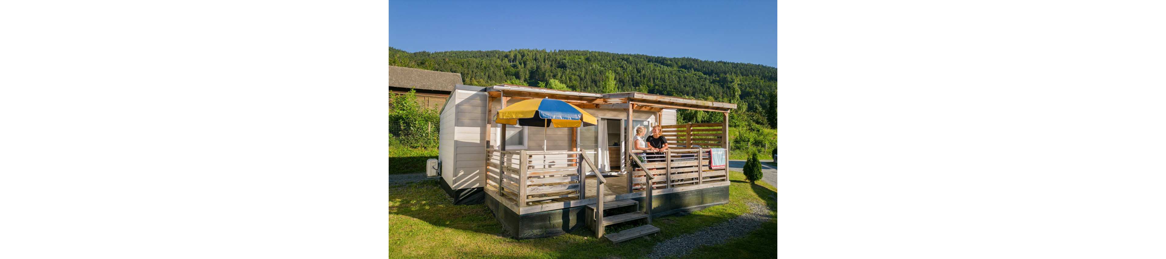 A couple stands on the wooden terrace of a modern mobile home set in a lush green landscape. The structure features a covered deck with railings, steps, and a colorful patio umbrella. Bright sunlight and clear blue skies highlight the surrounding forested hills and grassy campsite. The scene conveys a relaxed vacation atmosphere in a rural outdoor setting.