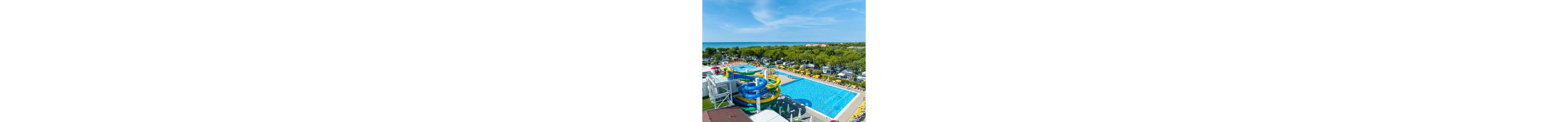 A large outdoor swimming pool with bright blue water is surrounded by yellow lounge chairs at a coastal resort. Colorful spiral waterslides in blue, yellow, and green dominate one side of the pool area. Beyond the pool, a dense line of green trees separates the resort from the distant beach and calm sea. The scene is captured on a clear, sunny day with a bright blue sky and scattered clouds.