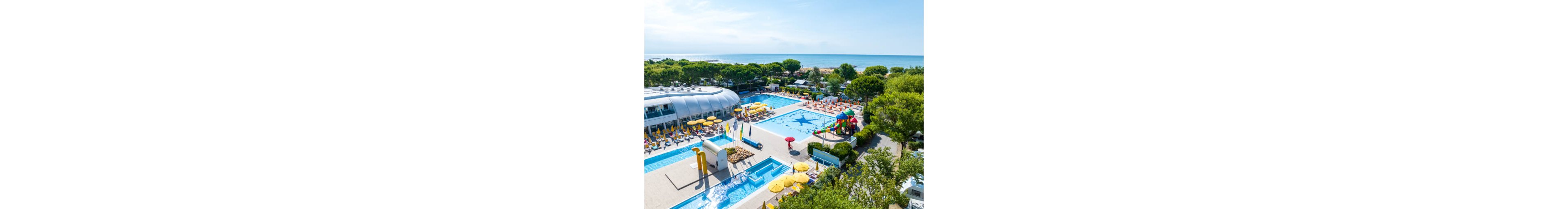 A large outdoor resort pool complex is shown near a sandy beachfront and calm blue sea. Multiple swimming pools with bright blue water are surrounded by yellow sun loungers and umbrellas. A colorful children’s water play area with slides and structures stands beside the main pool. Lush green trees frame the scene, creating a relaxed holiday atmosphere under a clear sky.