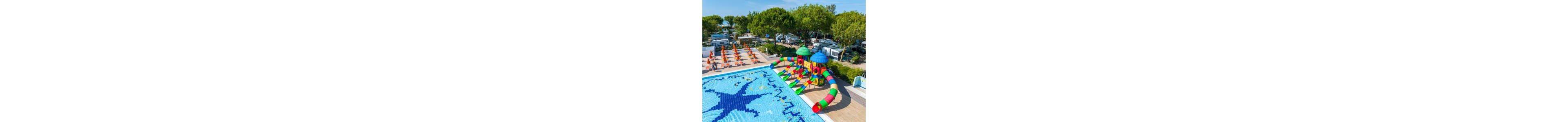 A vibrant outdoor swimming pool features a colorful multi-slide water playground shaped like an octopus. The poolside area is lined with rows of orange sun loungers and umbrellas, suggesting a family-friendly holiday setting. In the background, numerous camper vans and tents are nestled among tall green trees, indicating a busy campsite or holiday park. The bright blue pool tiles form playful patterns, enhancing the fun and summery atmosphere.