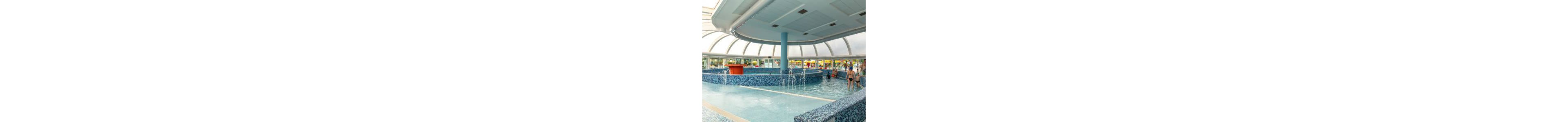 An indoor leisure swimming pool features a circular central basin with multiple small fountains spraying into shallow water. The pool is finished with blue mosaic tiles and surrounded by a curved seating edge. A large glass dome roof lets in natural light and offers views of the outdoor area. Several people in swimwear enjoy the water and walk around the pool, creating a relaxed, family-friendly atmosphere.